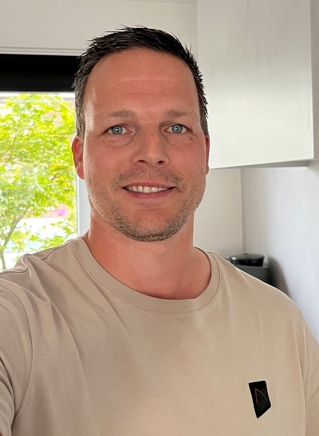 Een man met kort donker haar en blauwe ogen glimlacht in een beige T-shirt binnenshuis.