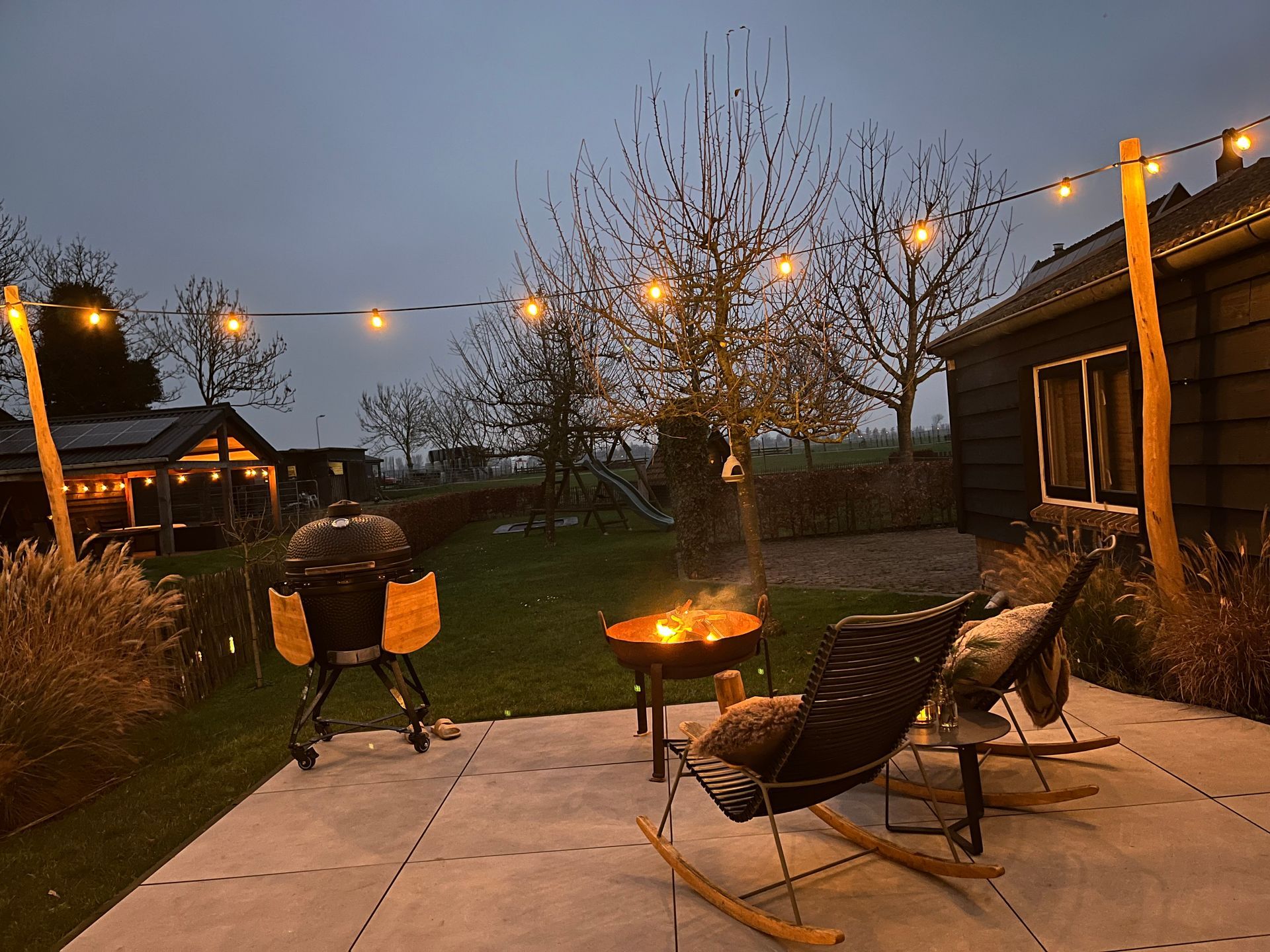 Buitenscène bij schemering met een vuurplaats, schommelstoelen en lichtslingers die een terras en een houten constructie verlichten.