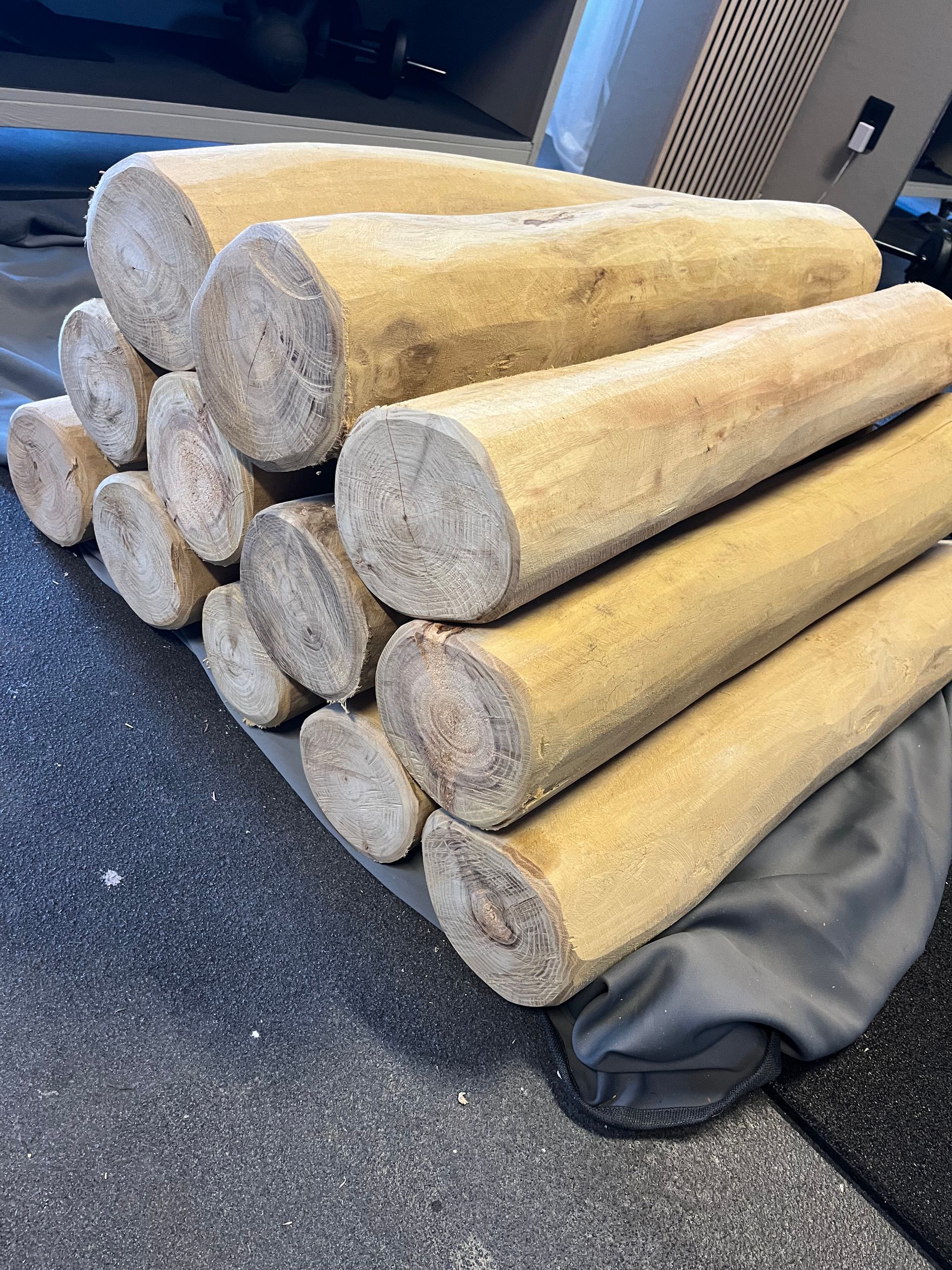 Stapel cilindrische houten balken van verschillende lengtes. Sommige hebben zichtbare kopse kanten. Op de achtergrond een grijze gymzaalvloer.