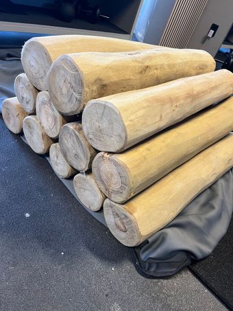Stapel cilindrische houten balken van verschillende lengtes. Sommige hebben zichtbare kopse kanten. Op de achtergrond een grijze gymzaalvloer.