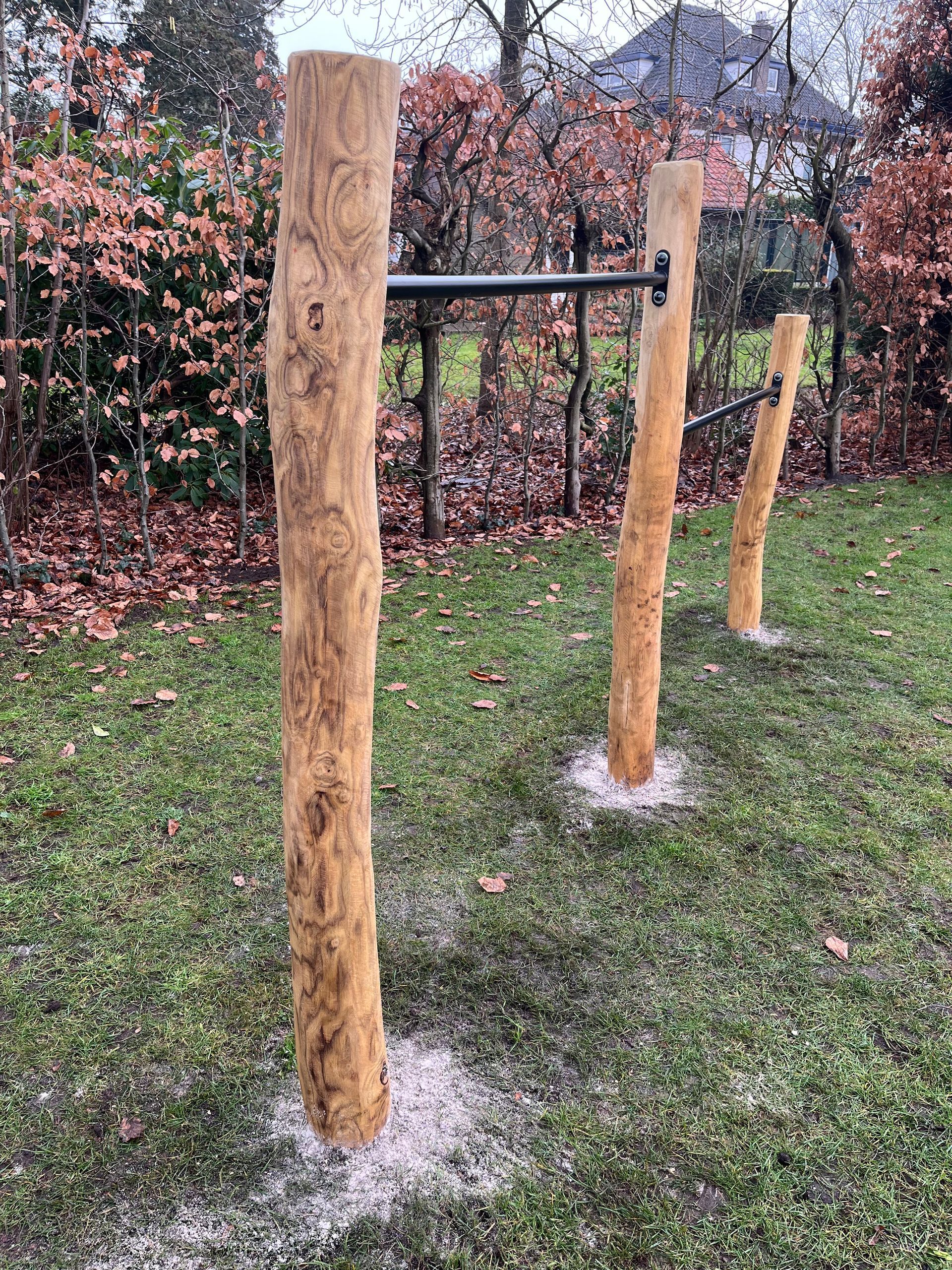 Optrekstang voor buiten met drie houten palen en een metalen stang. Geplaatst in een grasveld, omgeven door bomen.