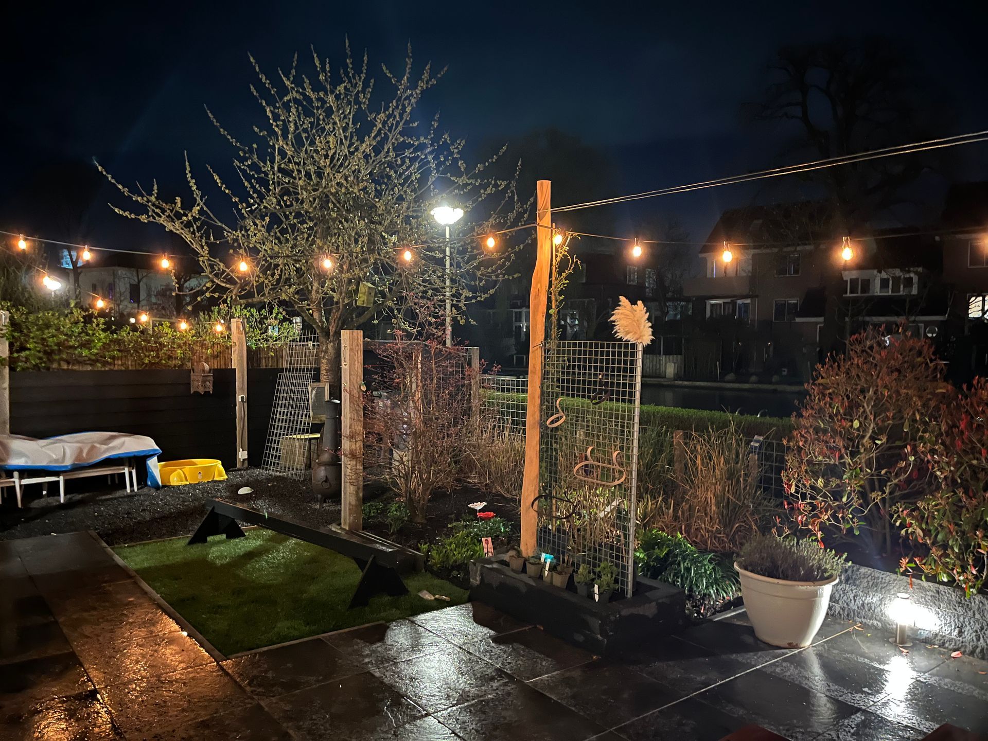 Buitenscène bij schemering met een vuurplaats, schommelstoelen en lichtslingers die een terras en een houten constructie verlichten.