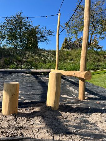 Stapel cilindrische houten balken van verschillende lengtes. Sommige hebben zichtbare kopse kanten. Op de achtergrond een grijze gymzaalvloer.