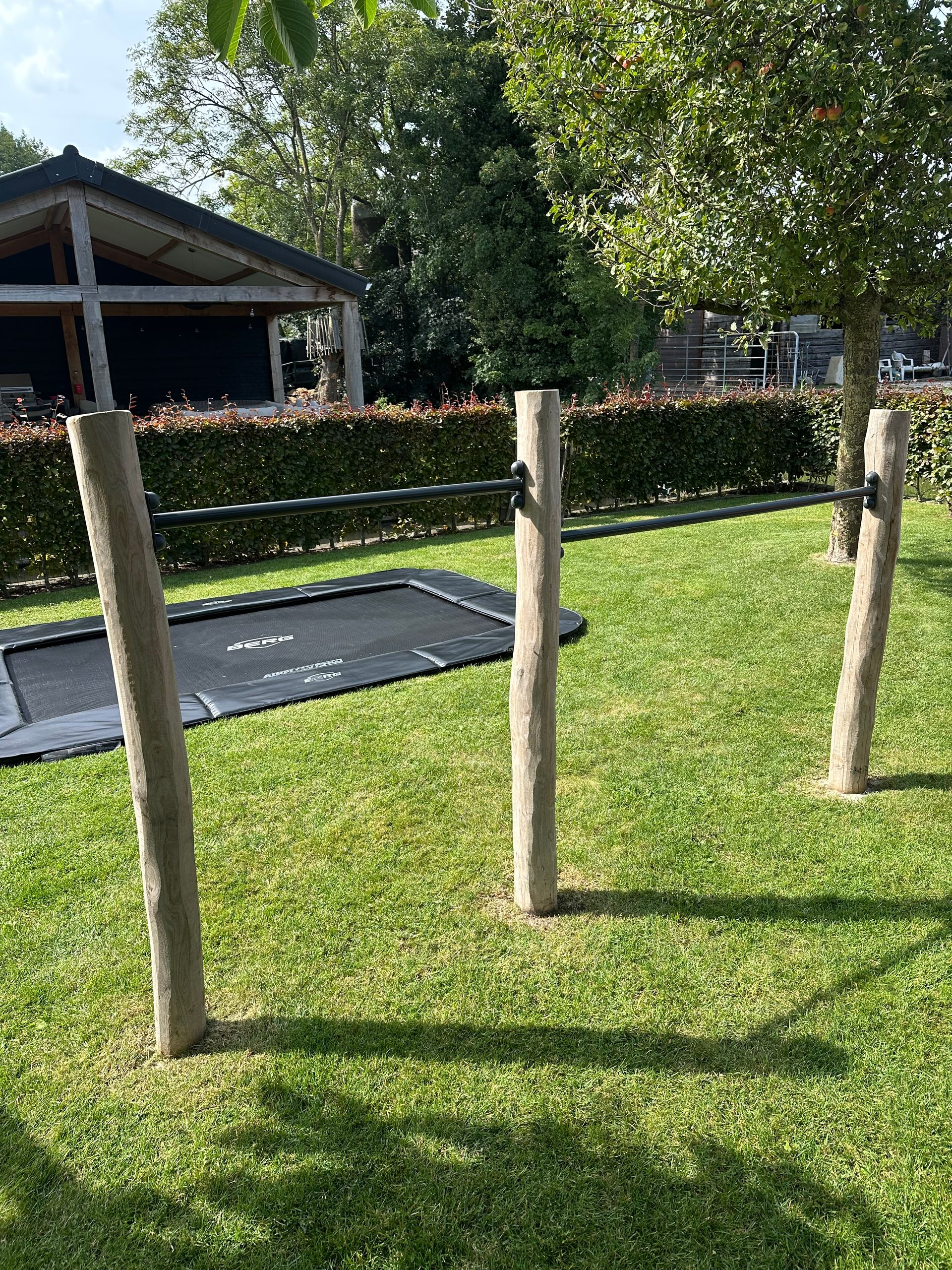 Drie houten palen met een horizontale dwarsbalk in een grasveld, met een trampoline en bomen.