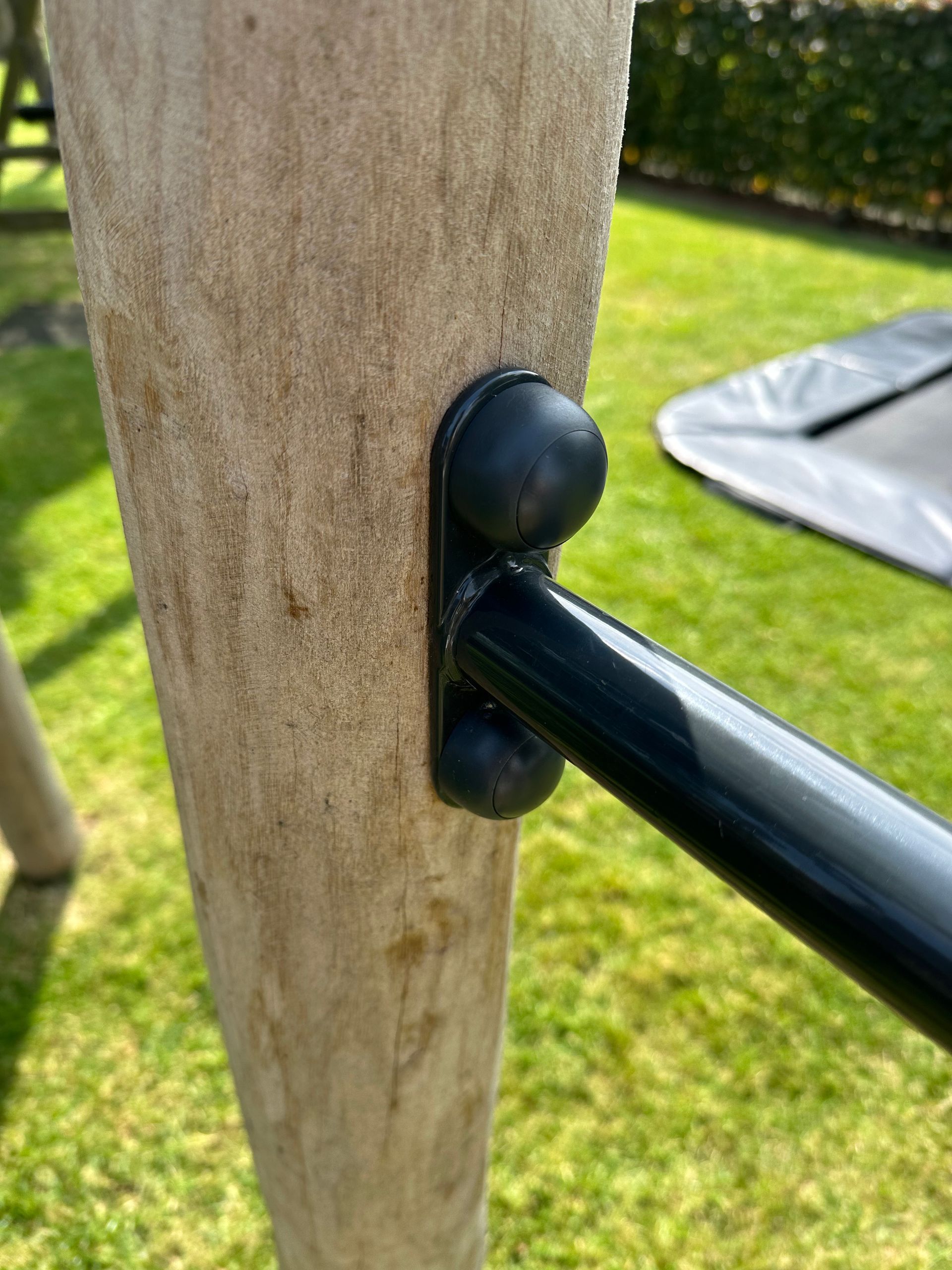 Zwarte stang bevestigd aan een houten paal met zwarte plastic fittingen in een grasrijke buitenomgeving.