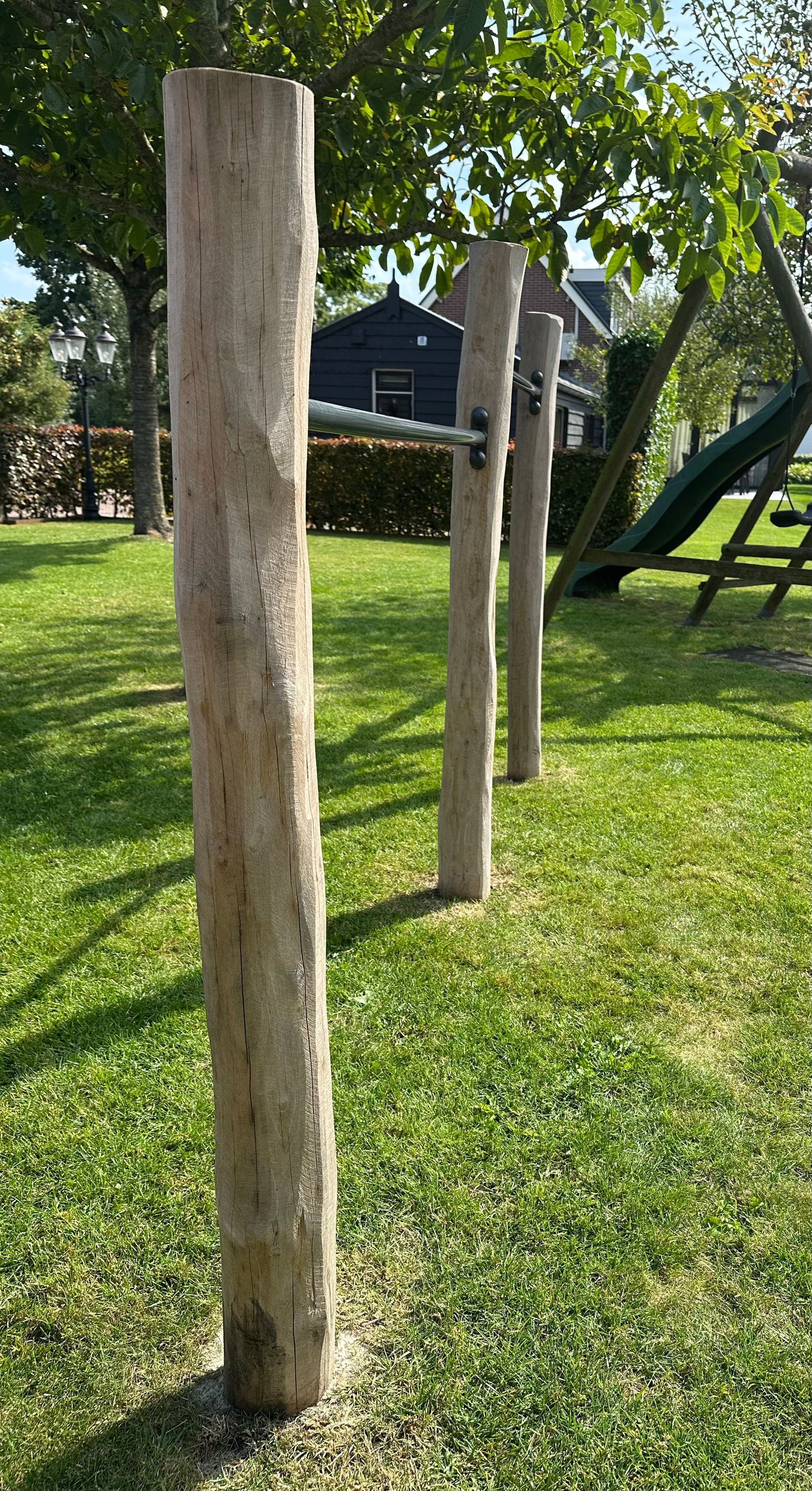 Drie houten palen in beton in een grasveld, met een grote boom en struiken op de achtergrond.