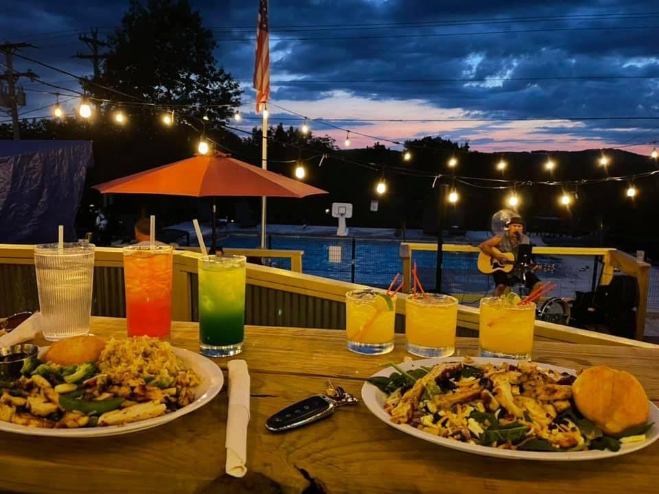 Outdoor dining scene at dusk; cocktails and food on a table overlook a pool, a musician plays.