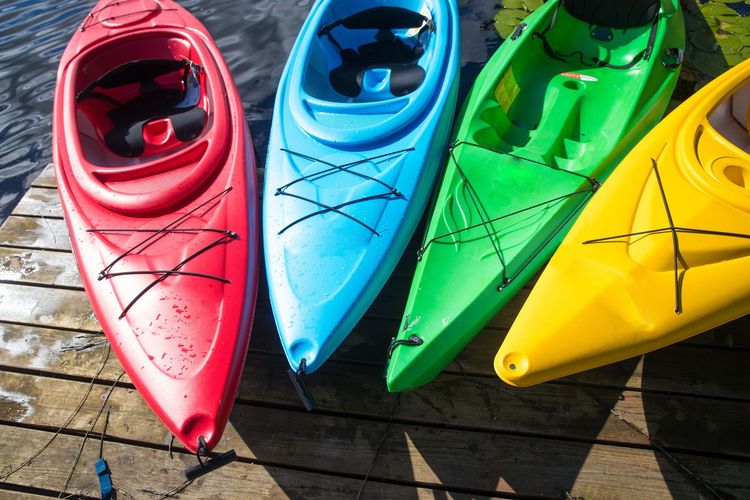 Four colorful kayaks: red, blue, green, and yellow, lined up on a wooden dock in sunlight.