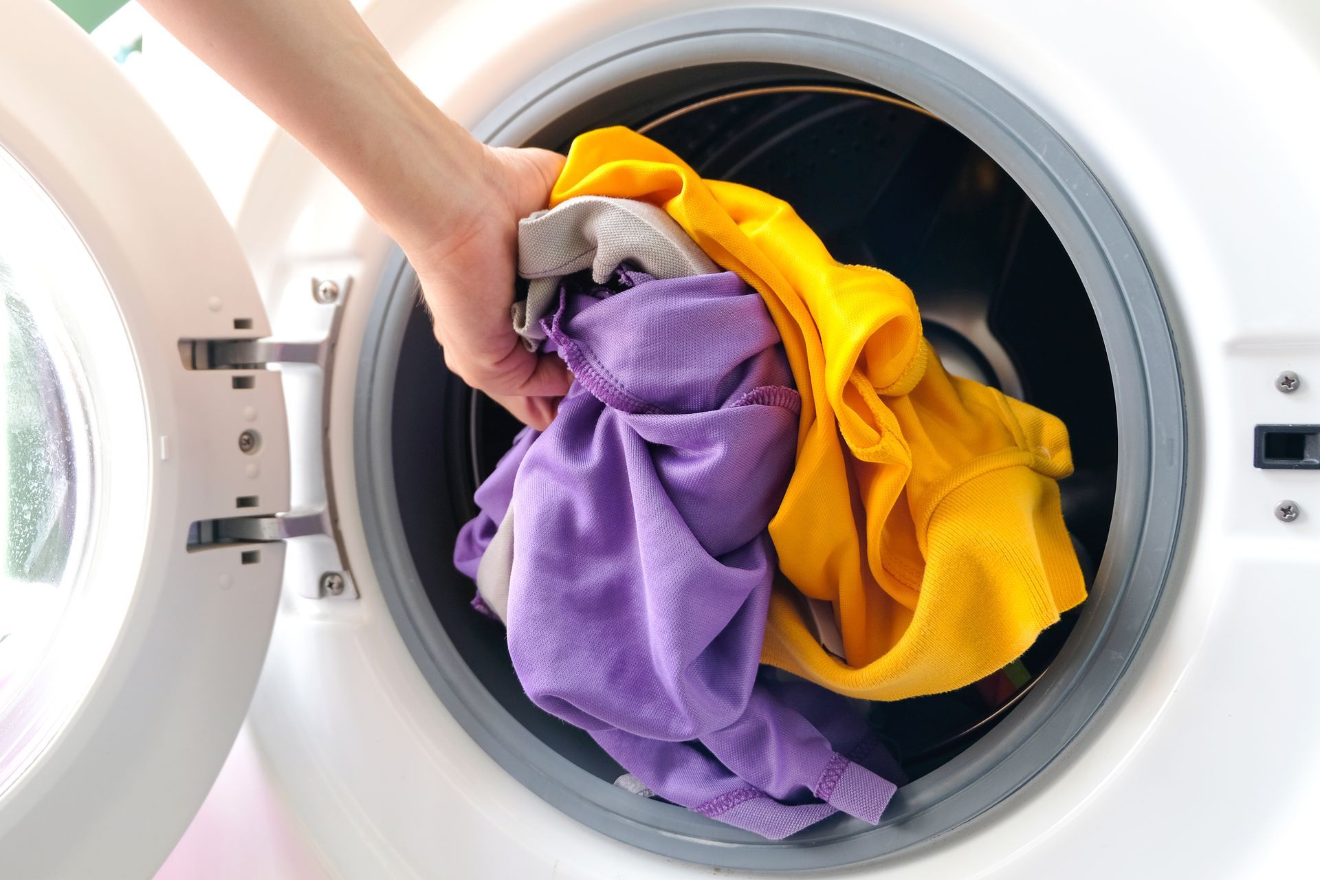 Hand placing colorful clothes into a washing machine.