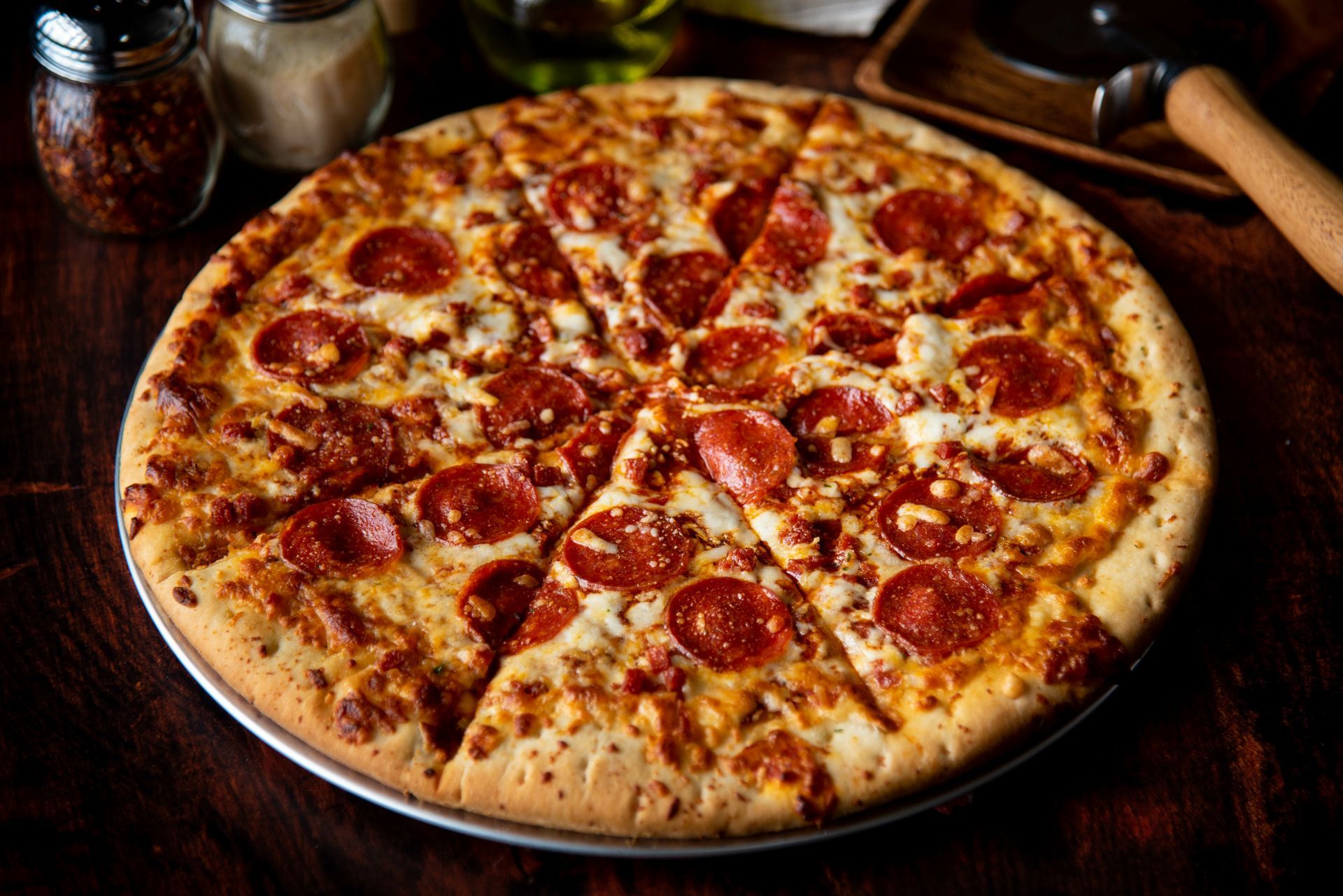 Pepperoni pizza, sliced into wedges, on a metal tray, with cheese, on a dark wooden table.