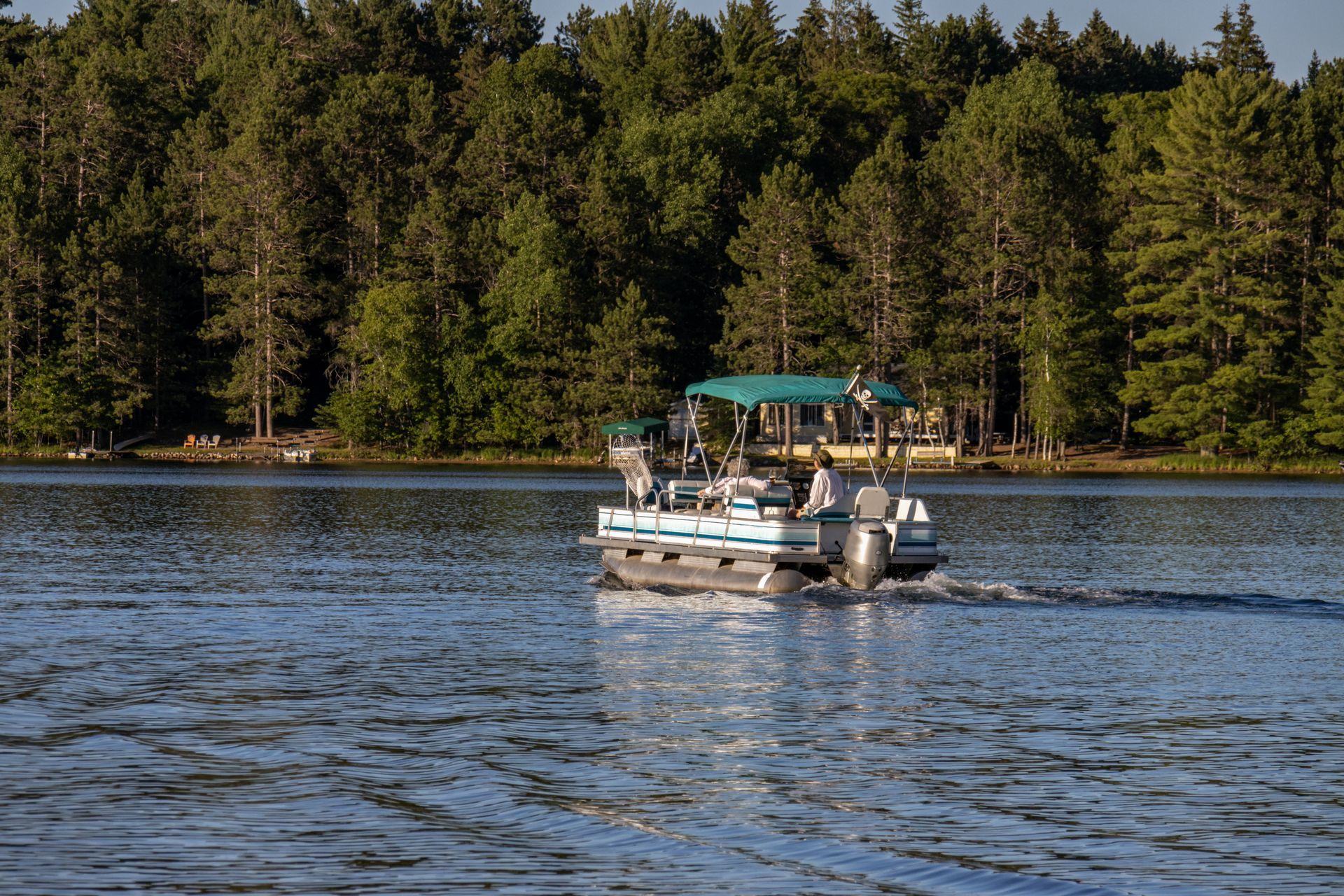 Pontoon boat on lake, moving toward shoreline with green trees.