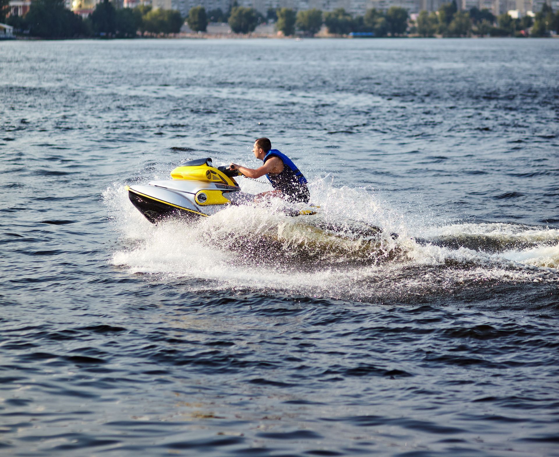 Man on a yellow jet ski creates a splash on a lake. He wears a life vest and faces forward.
