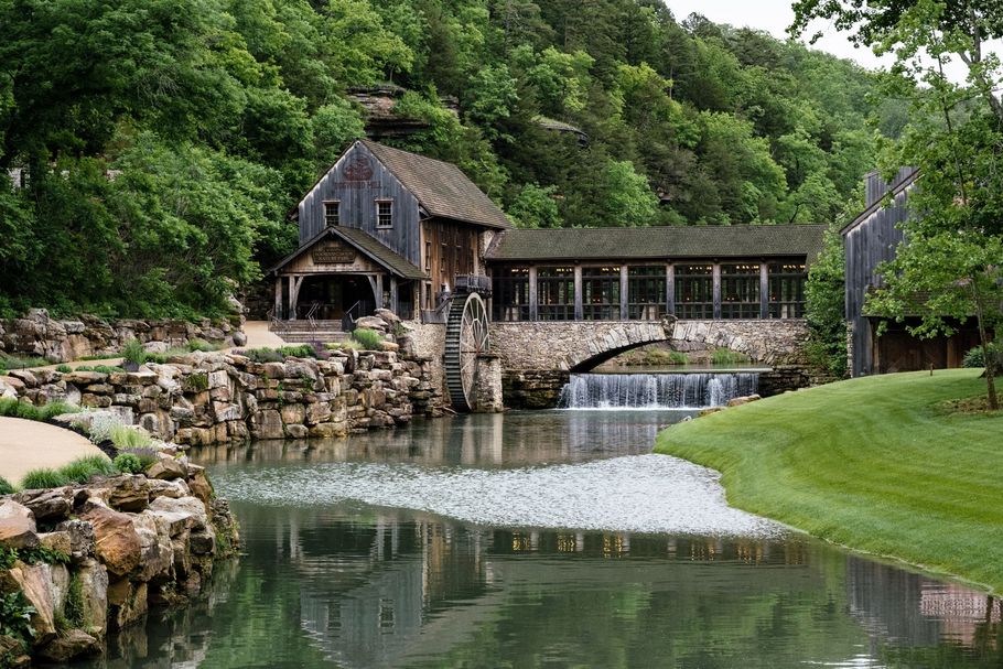 Old mill house and covered bridge over a creek with cascading water, surrounded by lush green trees.