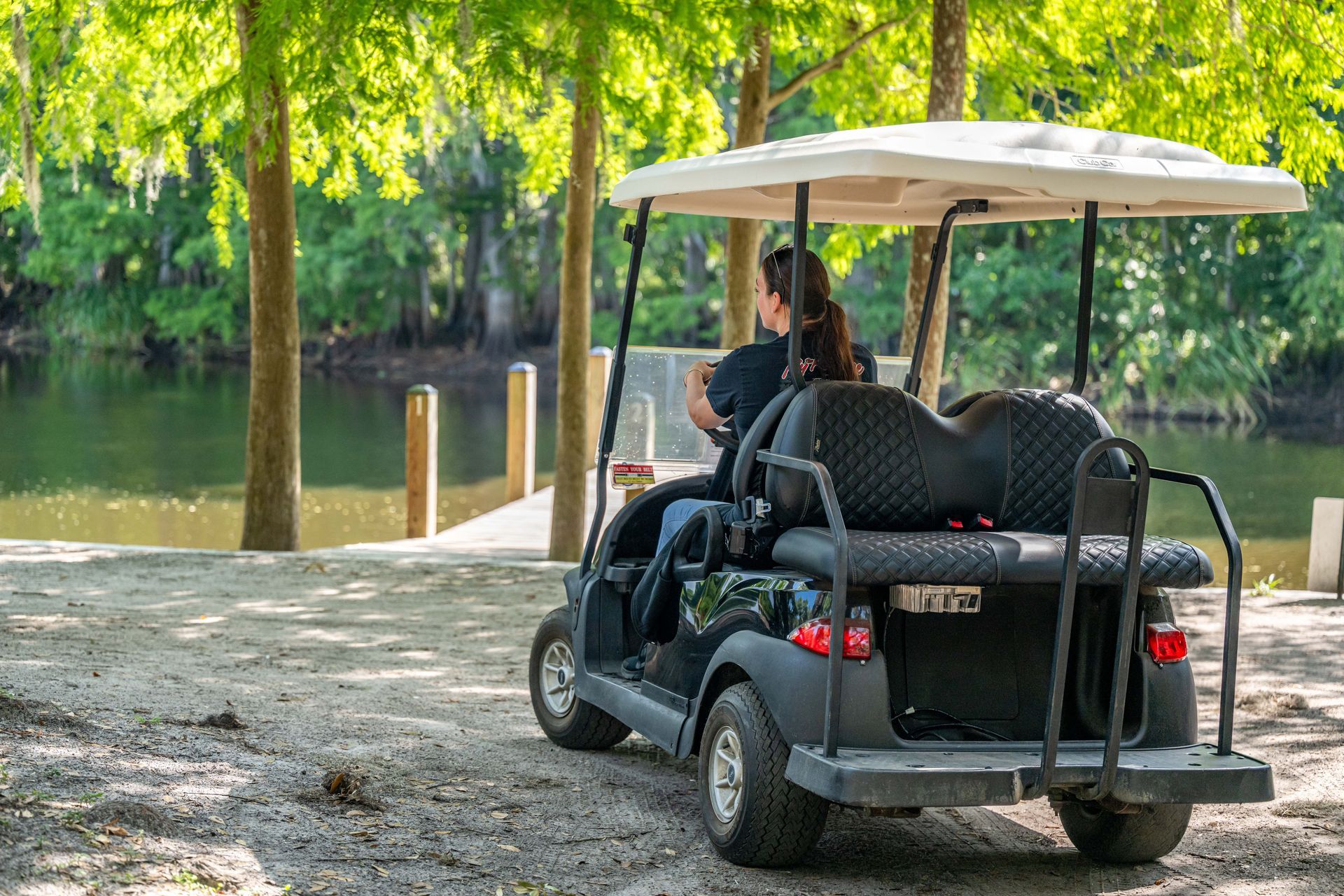 A person driving a black golf cart near a body of water, trees in the background.