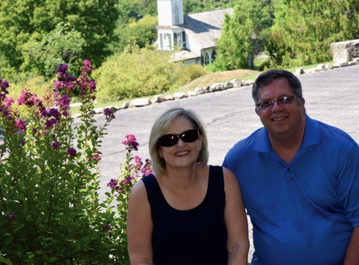 Woman with sunglasses and man smiling, posing in front of a building with white steeple, near purple flowers.