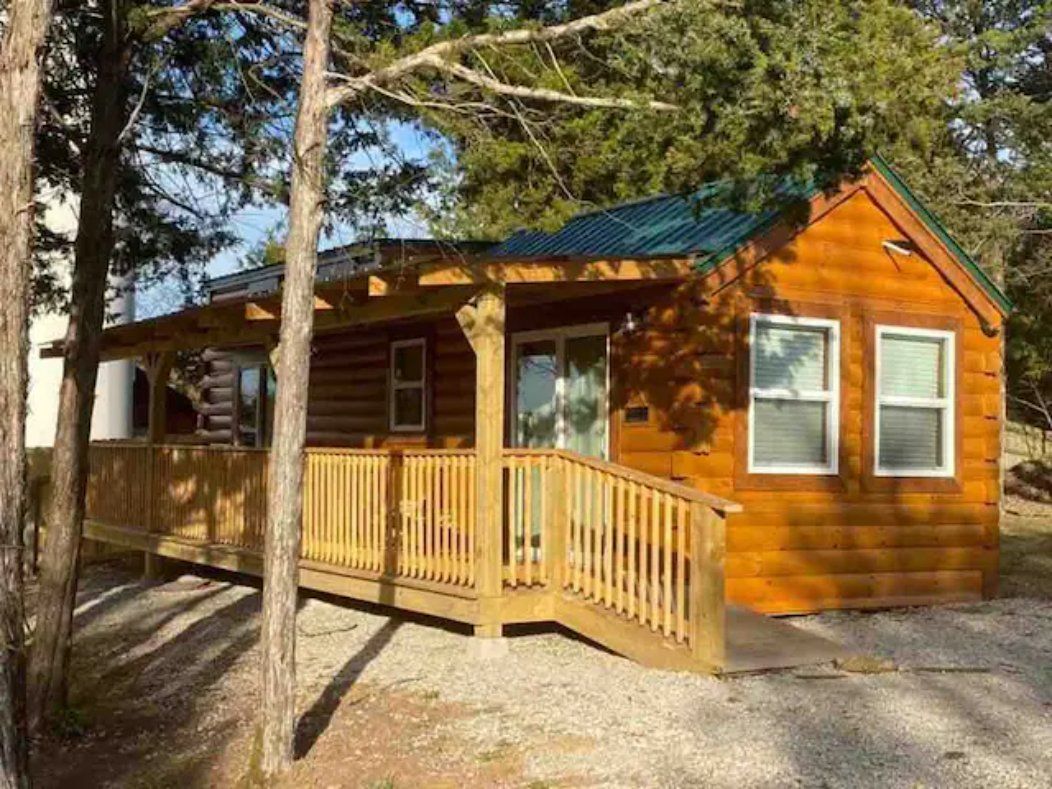 Log cabin with a wooden deck and ramp, nestled in a wooded area. The cabin has white-framed windows and a green metal roof.
