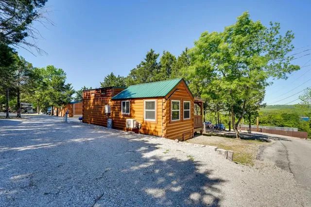 Wooden cabin with green roof, set on a gravel driveway, trees in the background under blue sky.