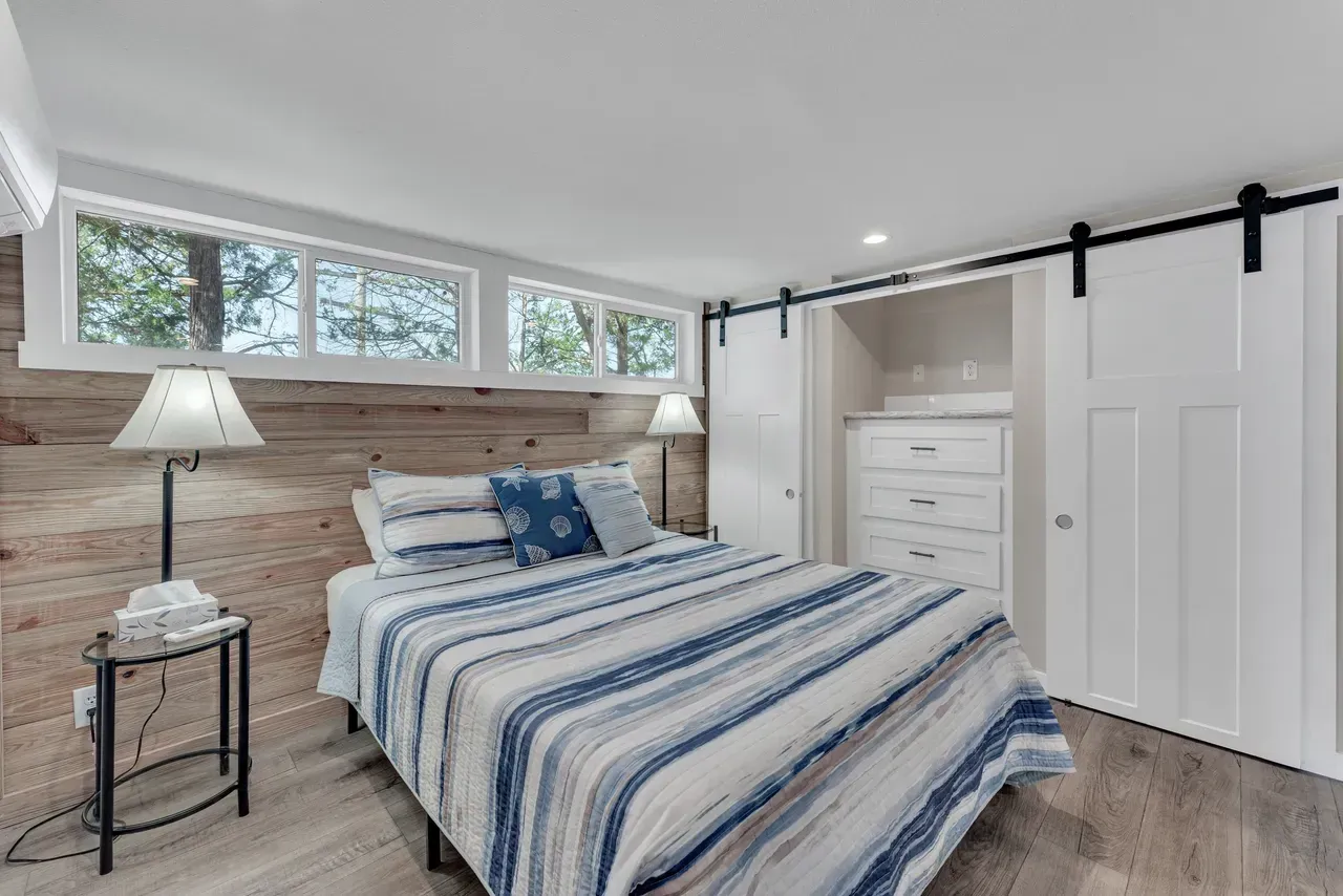 Bedroom with a bed, windows, lamps, and a closet with sliding doors. The walls are light wood.