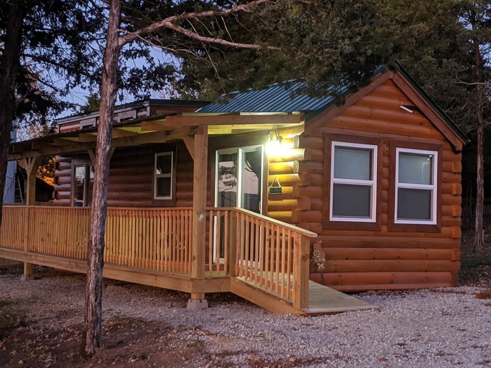 Log cabin with wooden porch and ramp, lit by a porch light, surrounded by trees.