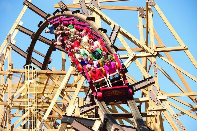 Roller coaster train with riders on a wooden track, ascending a curve against a blue sky.