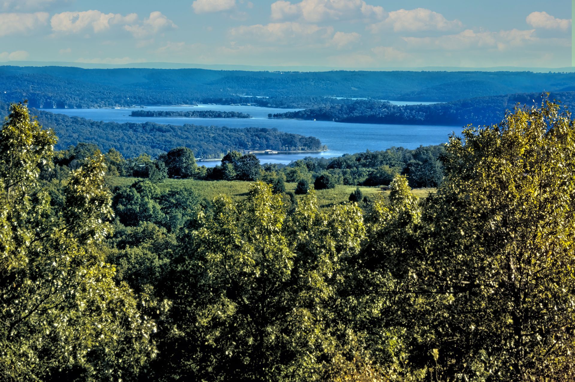 A scenic landscape view of a calm blue lake surrounded by rolling green hills and lush forested foreground foliage.