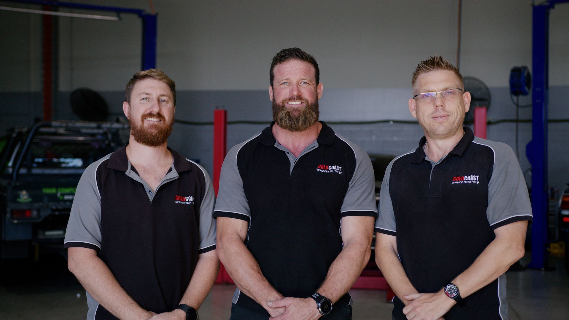 Three men in black and gray work shirts standing in a garage with a car in the background