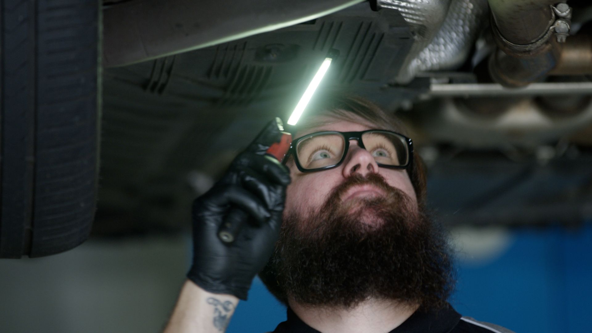 Mechanic inspecting underside of a vehicle with a glowing flashlight, wearing gloves and safety glasses.