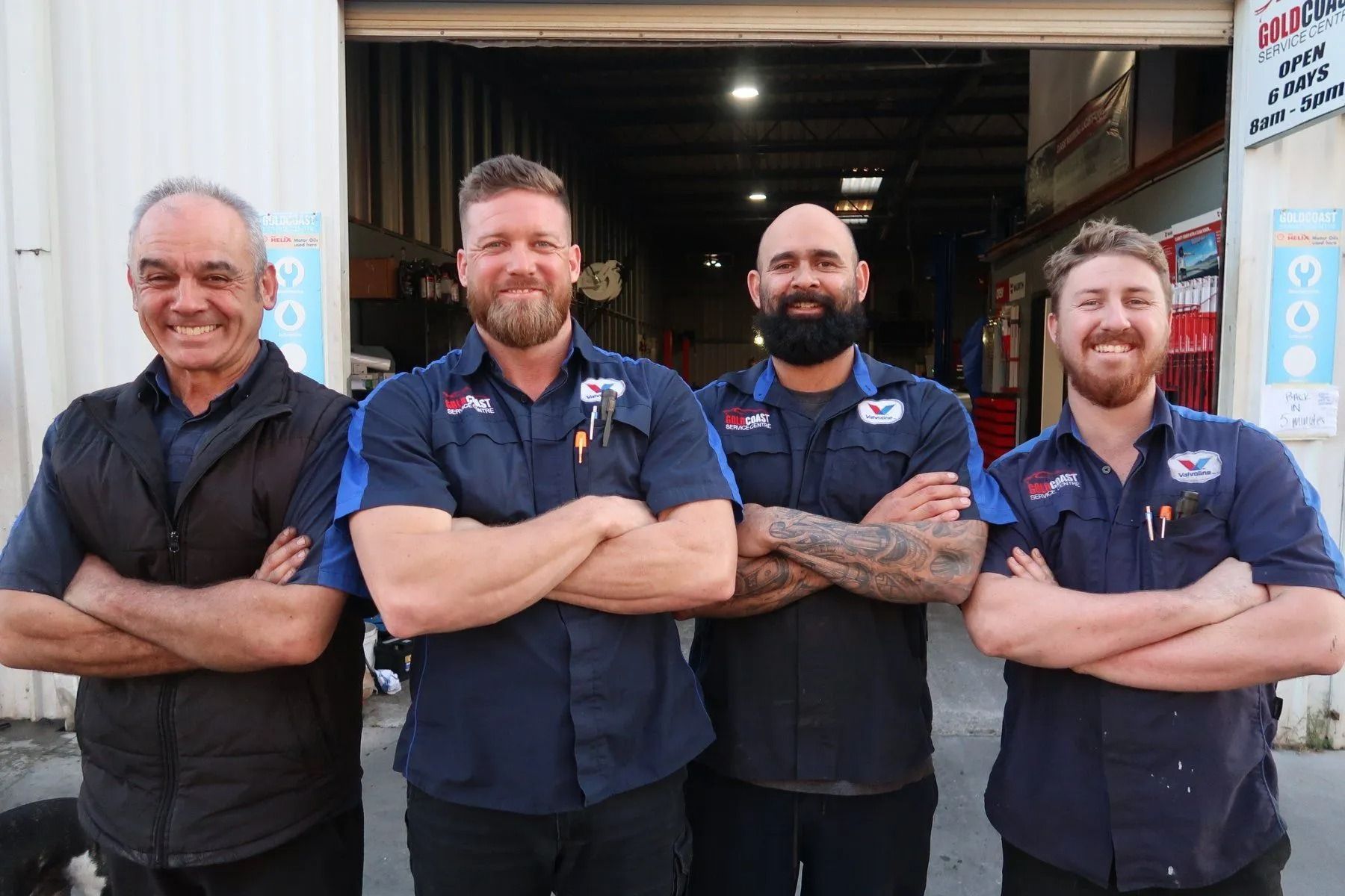 Four smiling people stand with arms crossed in front of a garage workshop, wearing matching navy work shirts.