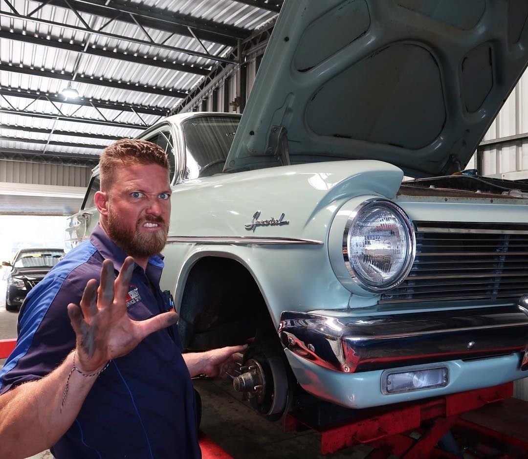 A mechanic with a dirty hand and an intense expression poses next to a light blue vintage car on a hydraulic lift.