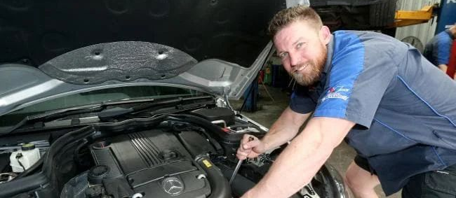 A mechanic smiles while working on the engine of a car inside a repair shop.