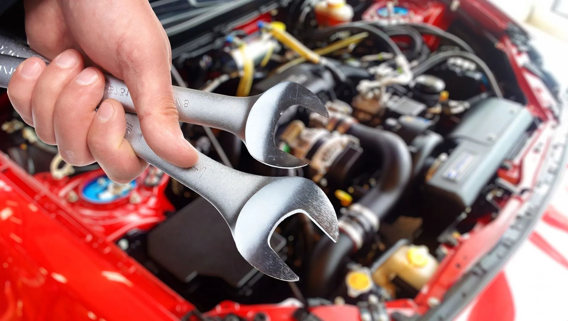 A hand holds two metal wrenches in front of the open hood and engine bay of a red car.