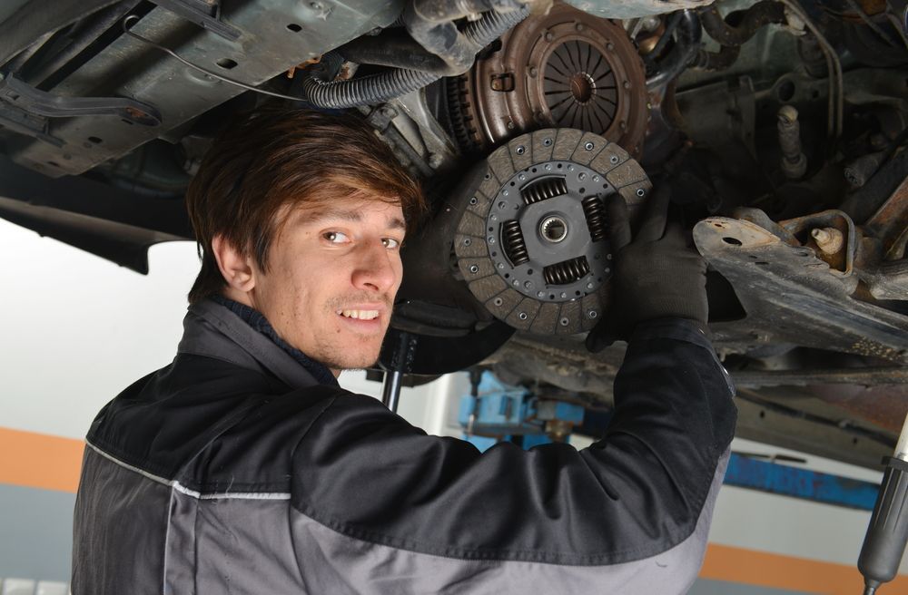A mechanic in a blue uniform writes on a clipboard while standing next to a car with its hood open in a repair shop.