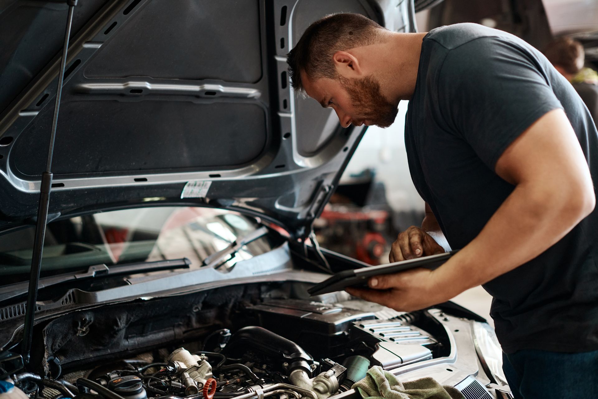 A mechanic in a garage reviews digital diagnostics on a tablet while inspecting an open car engine.
