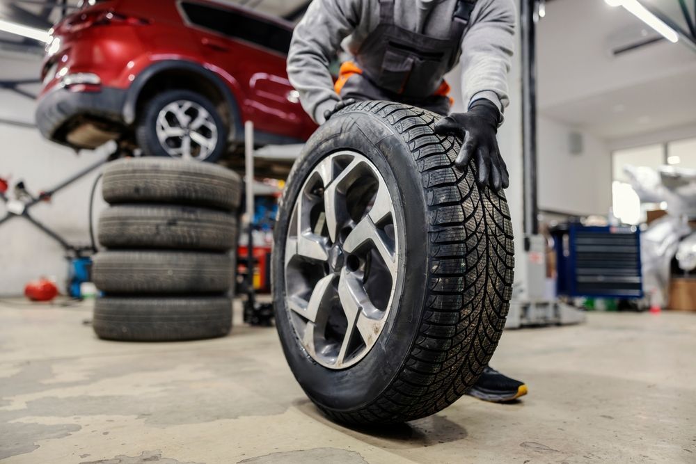 A mechanic in a garage holds an upright car tire with a metallic rim, with a red car on a lift in the background.