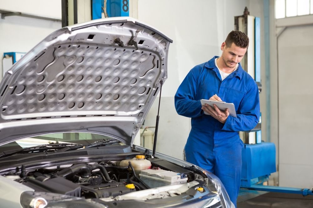 A mechanic in a blue uniform writes on a clipboard while standing next to a car with its hood open in a repair shop.