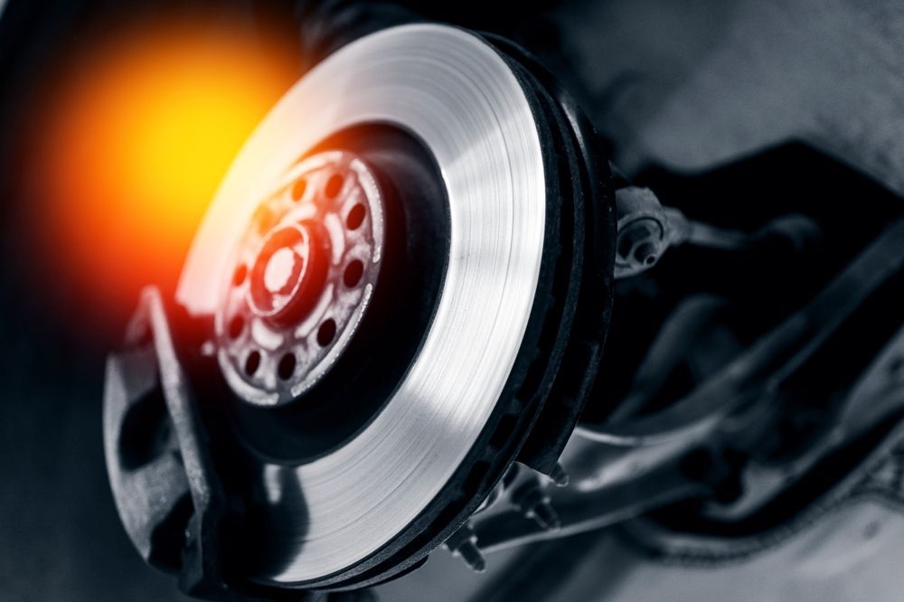A mechanic in a blue uniform writes on a clipboard while standing next to a car with its hood open in a repair shop.