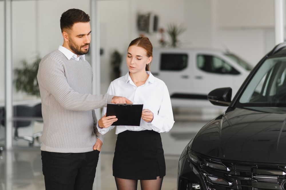 A person in a gray sweater points to a clipboard held by a professional in a white shirt and black skirt at a car dealership.
