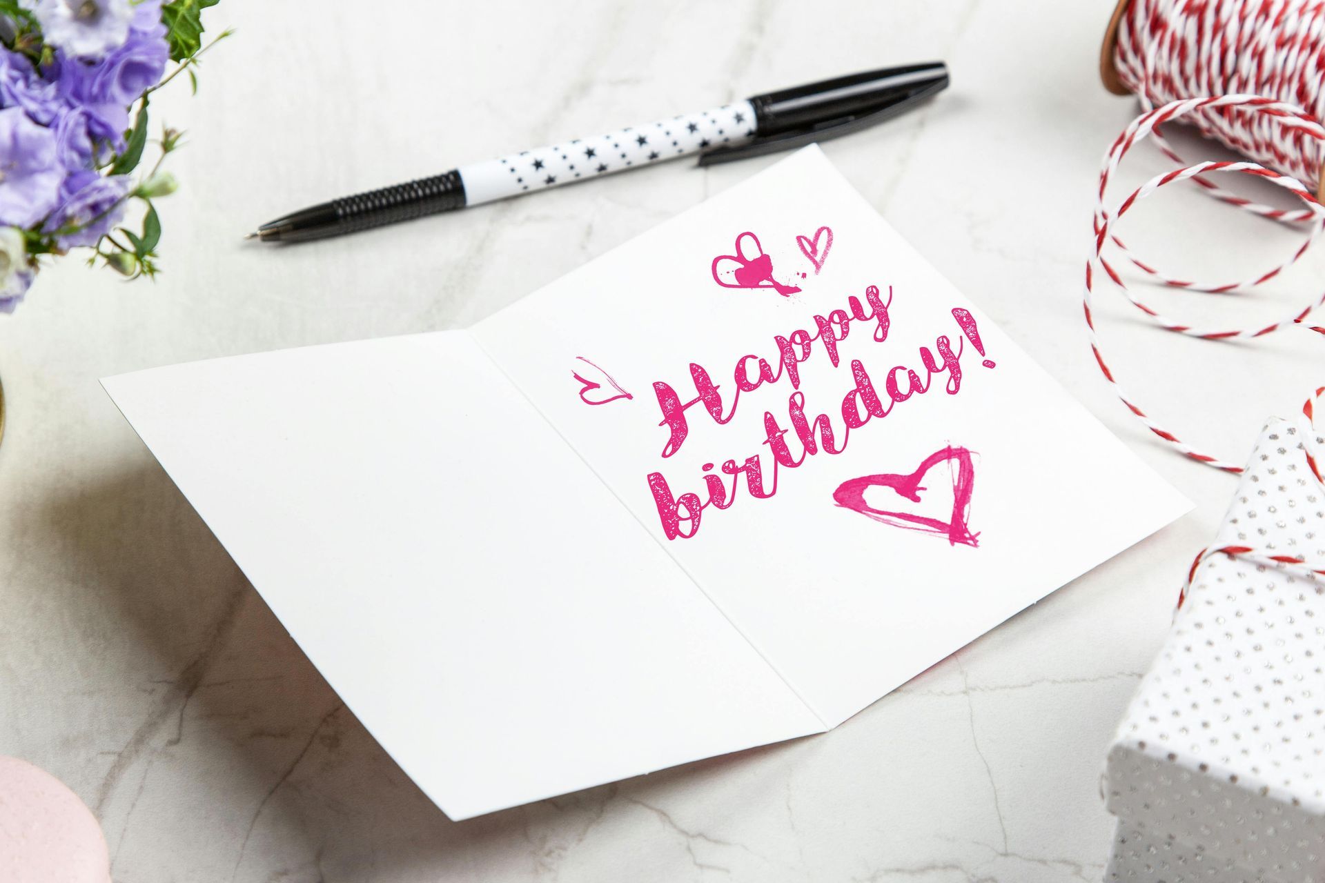 Birthday card with pink “Happy Birthday” lettering, pen, flowers, and gift wrap on a white desk