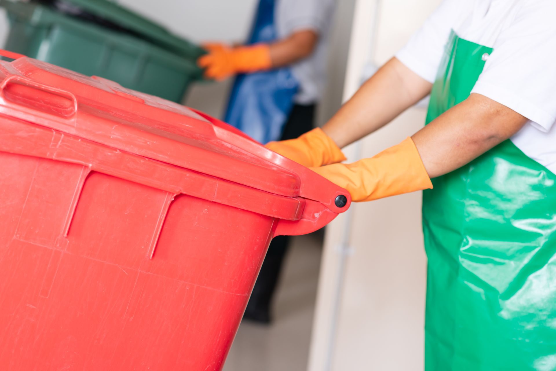 A partial view of two cleaners with gloves and aprons holding wheelie bins for cleaning.