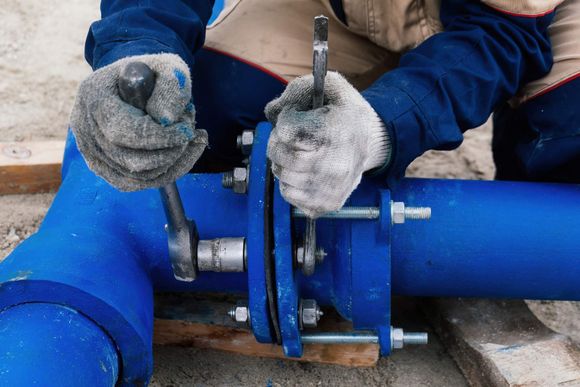 Person in gloves using wrenches to tighten bolts on a blue pipe flange outdoors.
