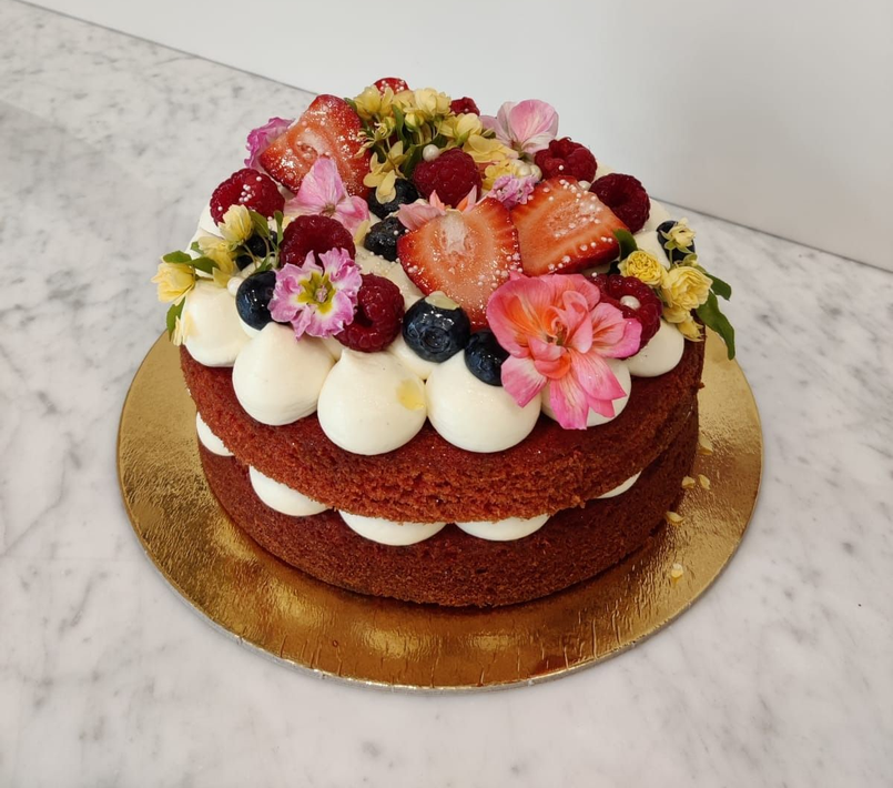 Torta a due piani senza copertura, con crema bianca, frutti di bosco e fiori su una base dorata.