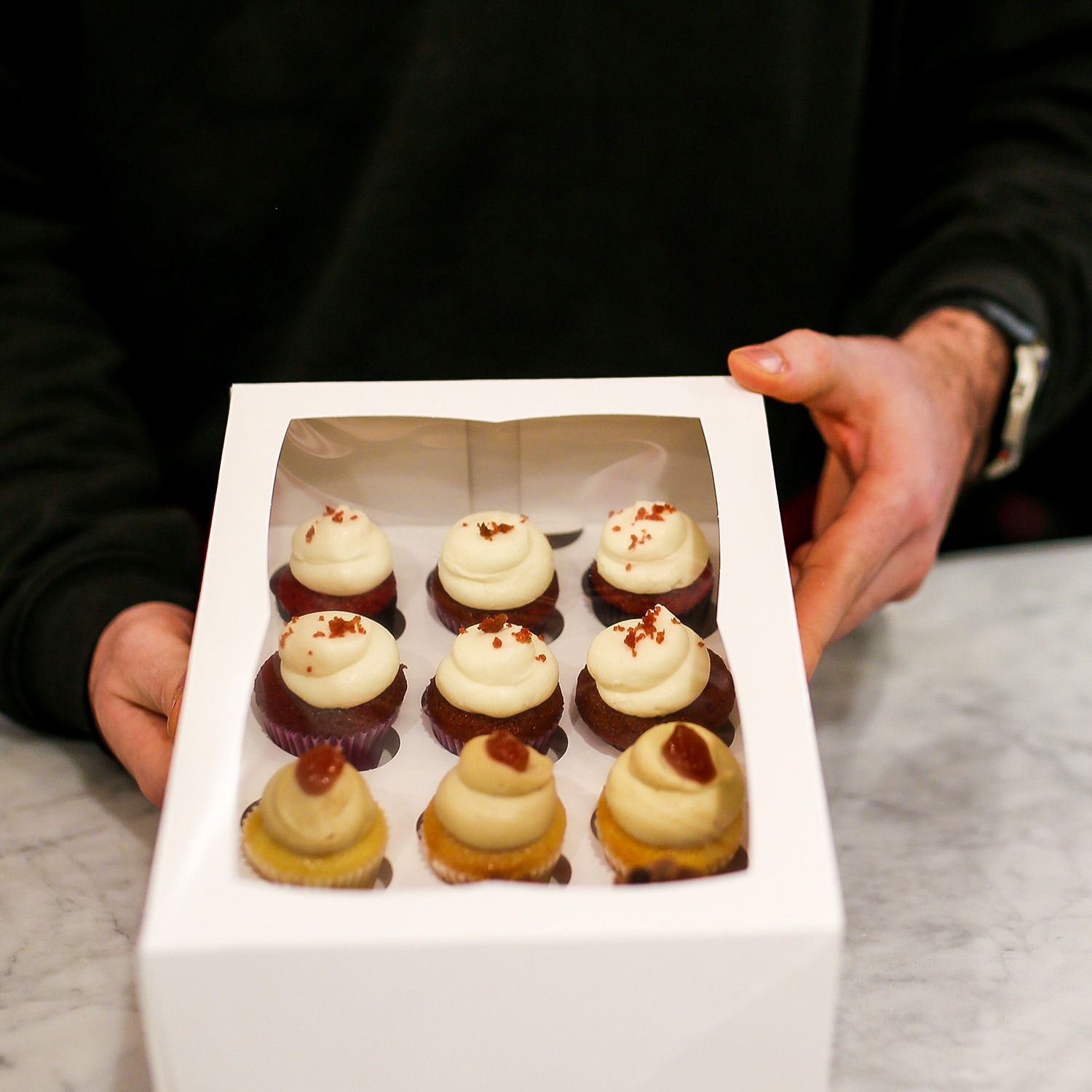 Scatola di mini cupcake assortiti con glassa bianca, tenuti in mano da qualcuno sopra un bancone di marmo.