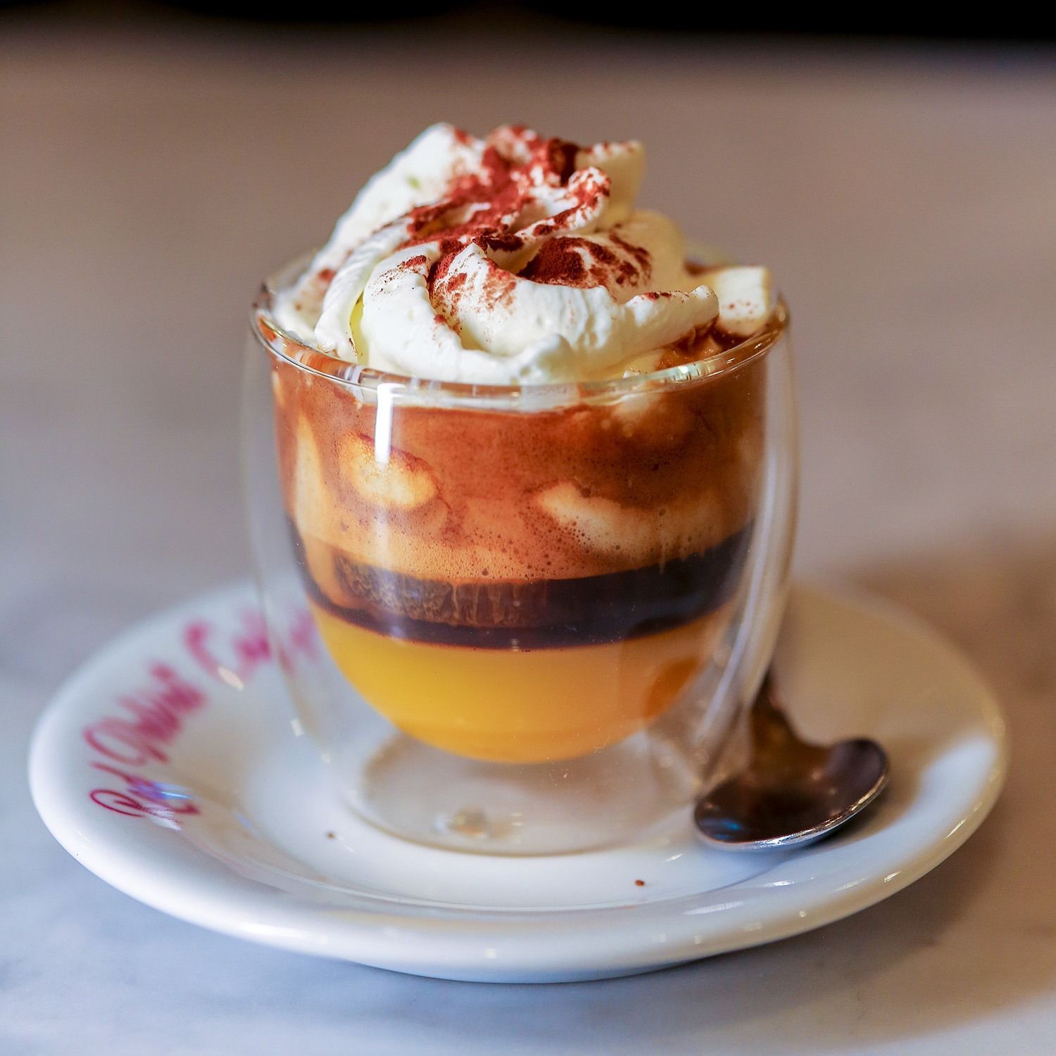 Dessert al caffè a strati in una tazza di vetro, guarnito con panna montata e una spruzzata di salsa rossa su un piattino.