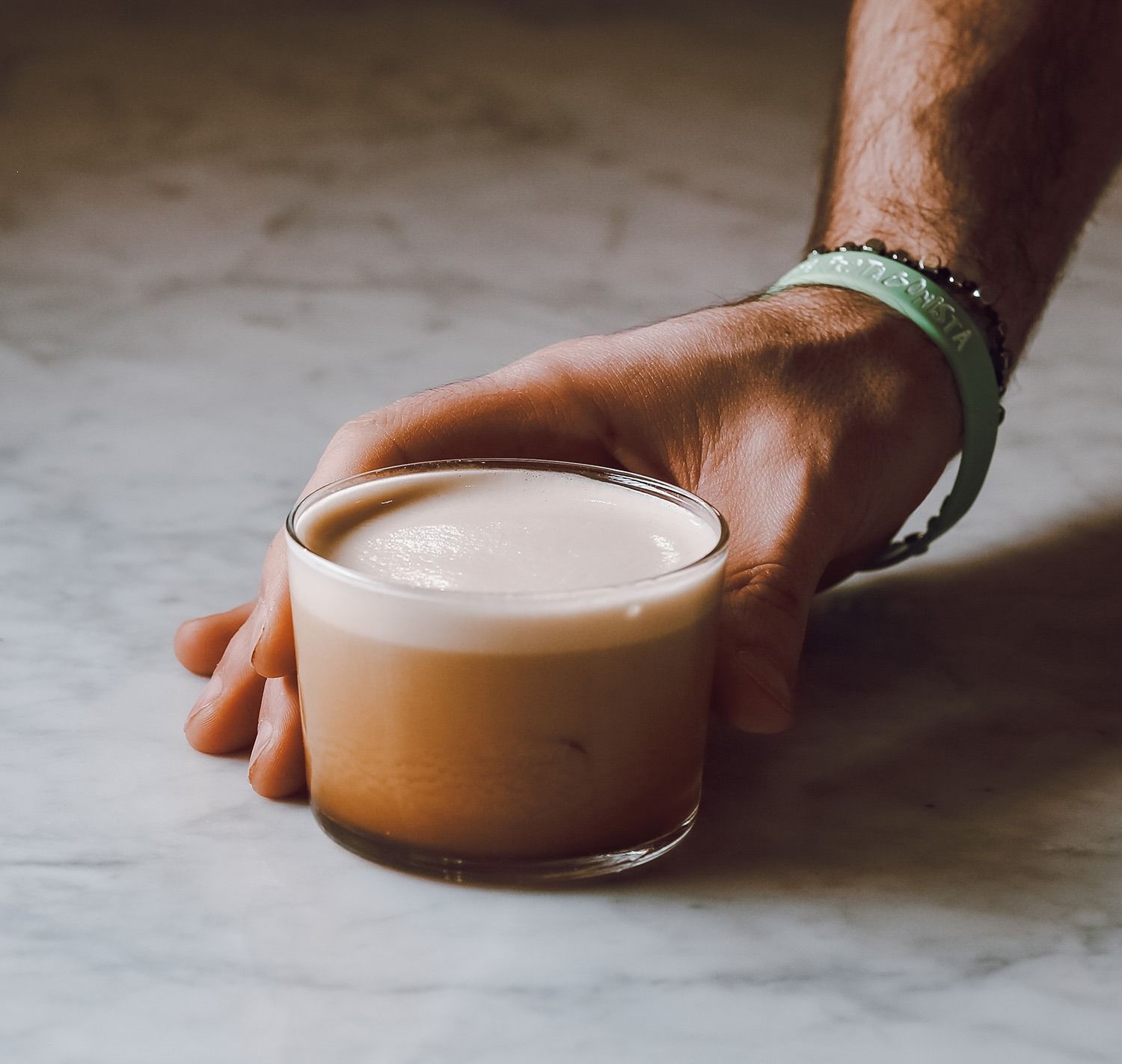 Una mano che regge una tazza di vetro trasparente con del caffè schiumoso su una superficie di marmo.