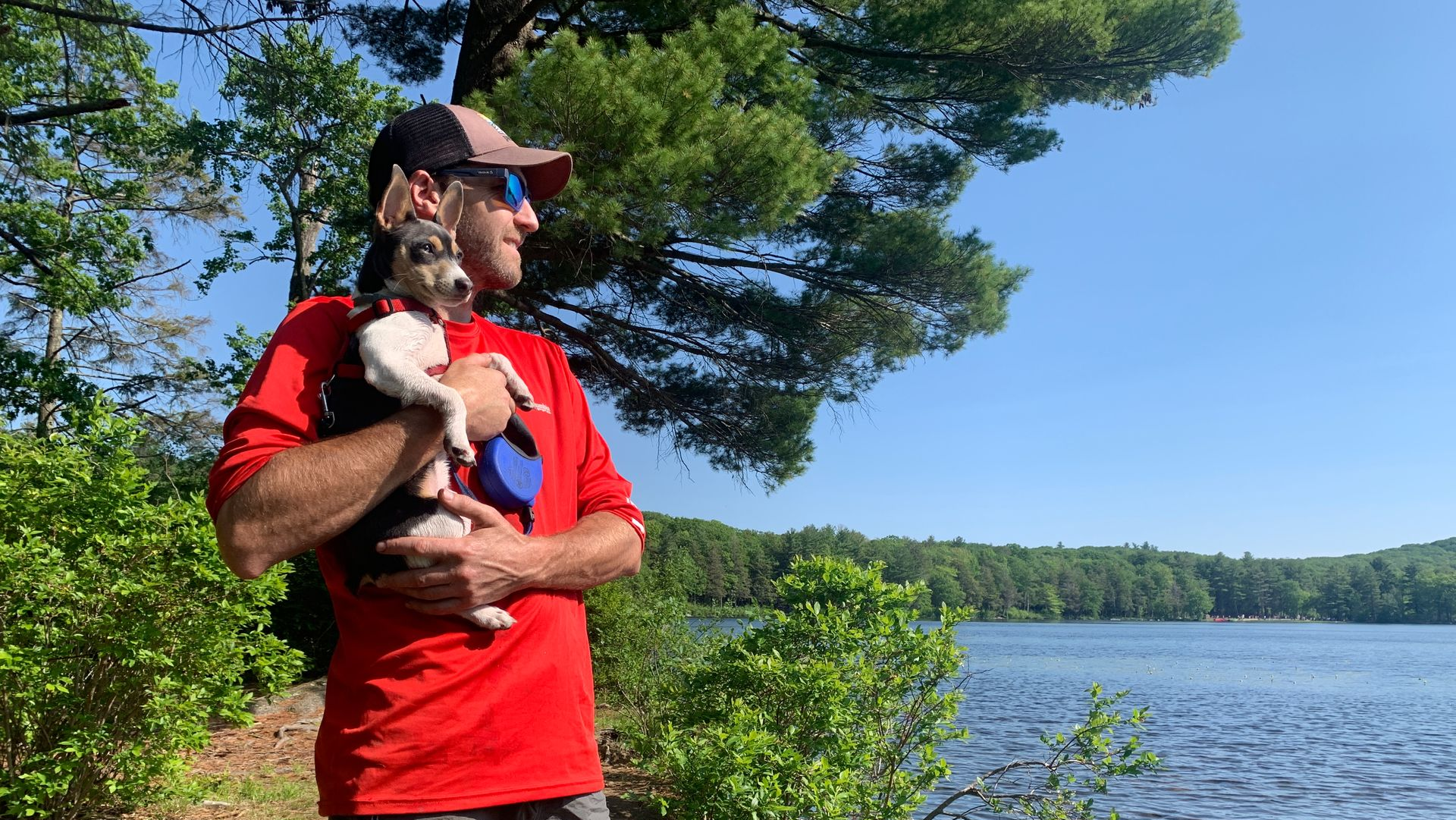 Man holding dog by a lake, looking at the water. Both looking away from camera, sunny day, blue sky.