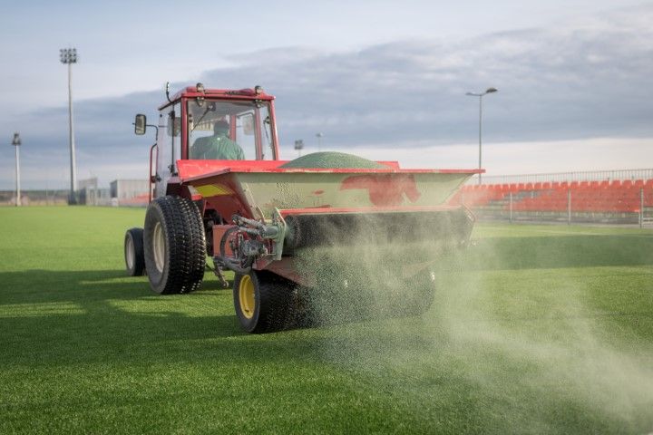 Tractor spreading green fertilizer on a soccer field, with a person in the cab, on a cloudy day.