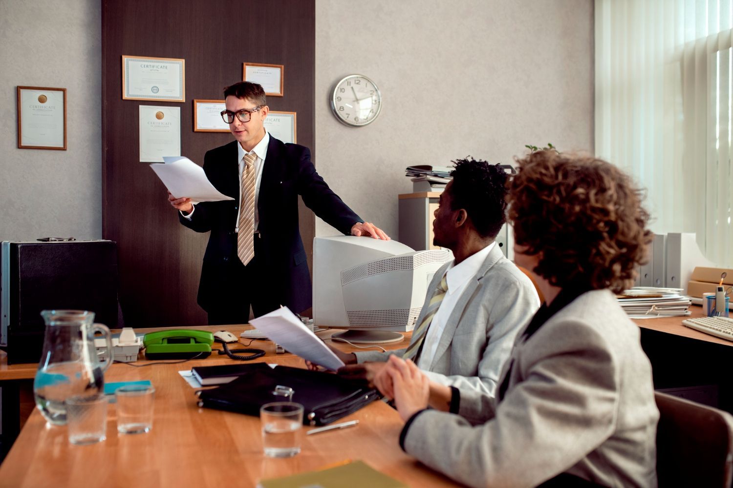 A person in a suit stands in an office meeting, presenting papers to two colleagues seated at a wooden table.