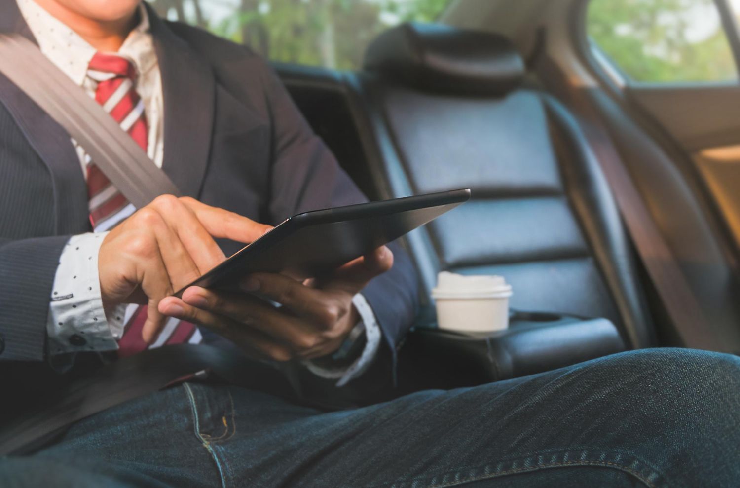 A person in a suit and tie sits in the back of a car, using a tablet while a cup sits in the center console.