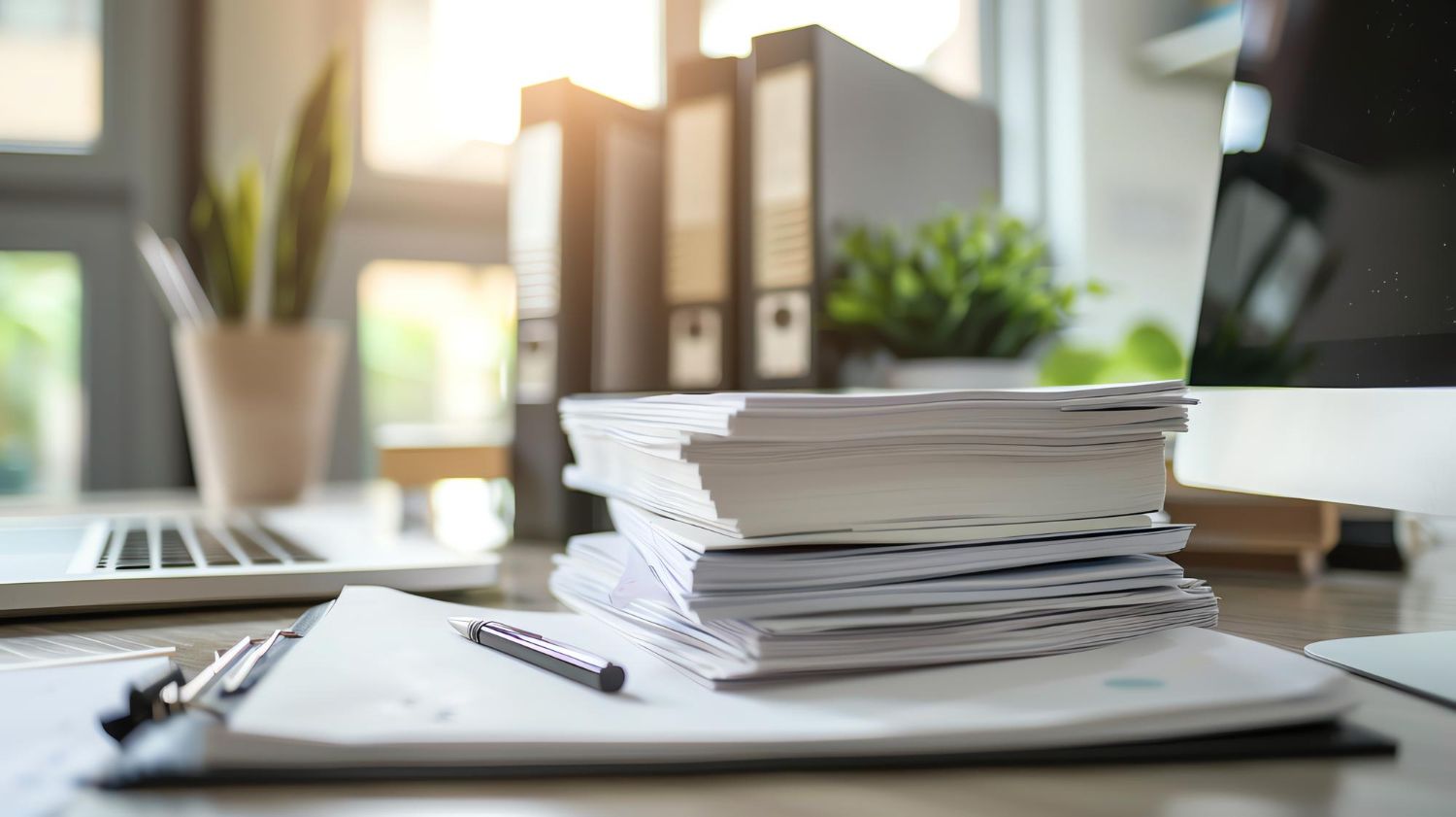 A cluttered office desk with a large stack of papers, a black pen, binders, a plant, and a laptop in a bright workspace.