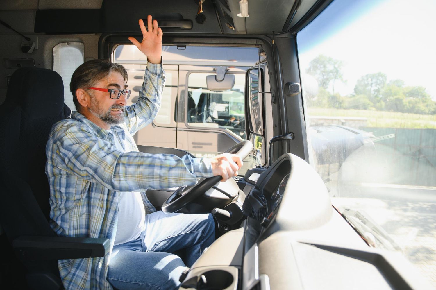 A person in a plaid shirt sits in a truck cab, holding the steering wheel with one hand and waving with the other.
